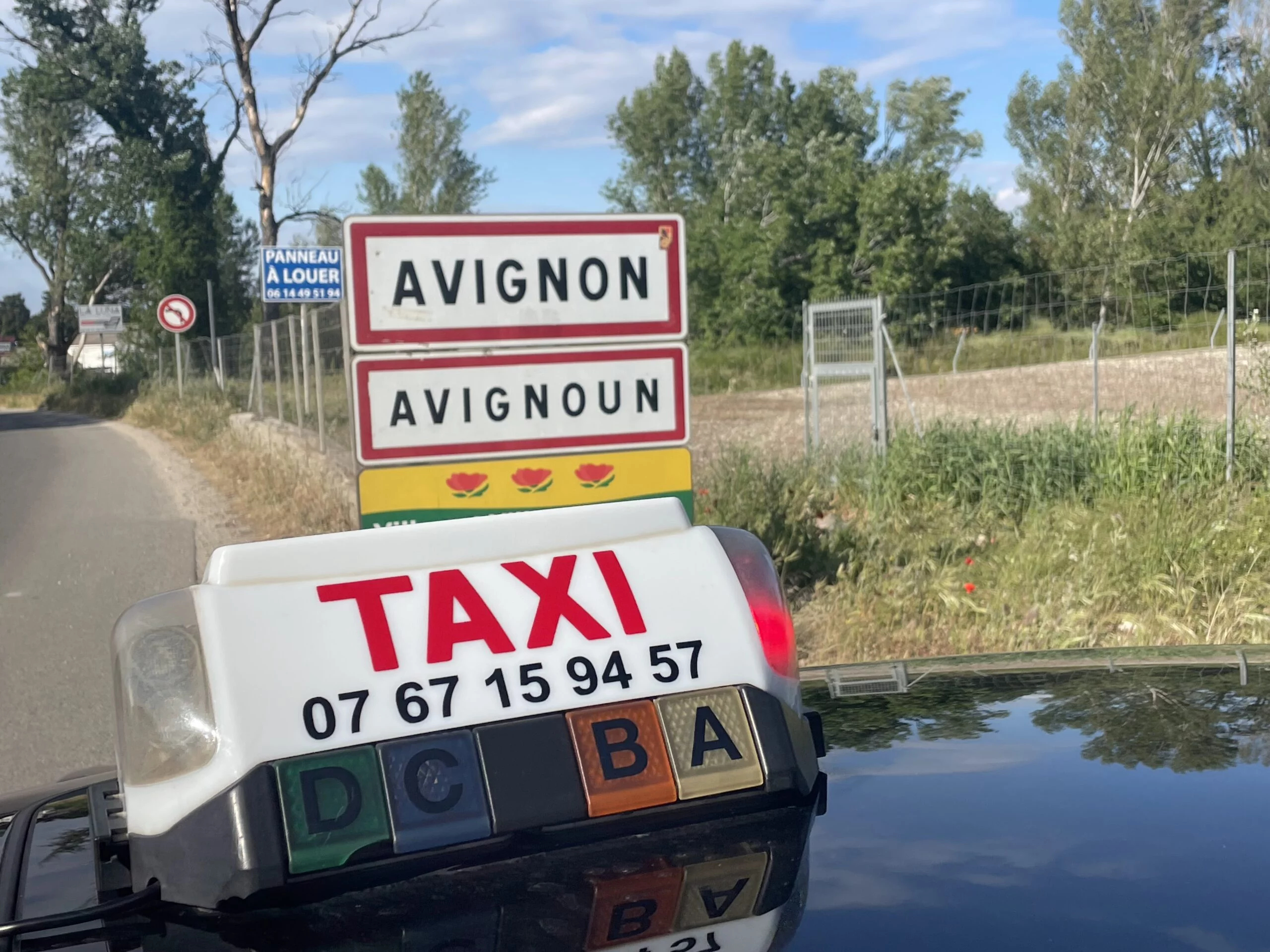 Photos d'un taxi devant le panneau d'entrée du village d'Avignon. On y voit l'enseigne du taxi et le panneau de la ville d'Avignon. Le taxi est sur une route. On voit également des champs avec des clôtures et des panneaux de signalisation.